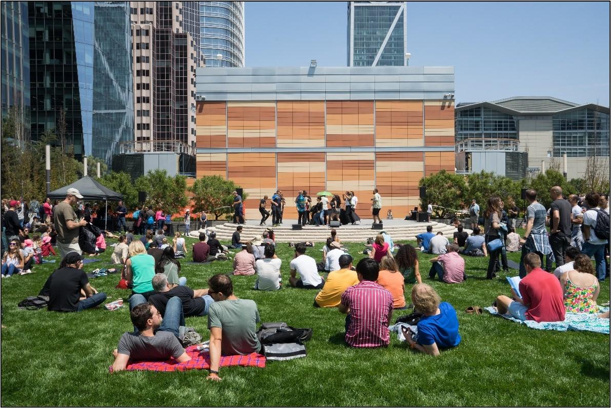 People sit on a lawn watching a performance on a stage.