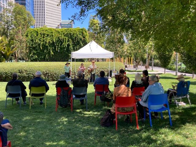 photo of crowd sitting on chairs on lawn in the shade, a tent with musicians is in the background.