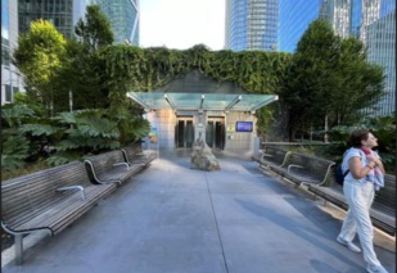 A small plaza with benches, surrounded by greenery and buildings in the background.